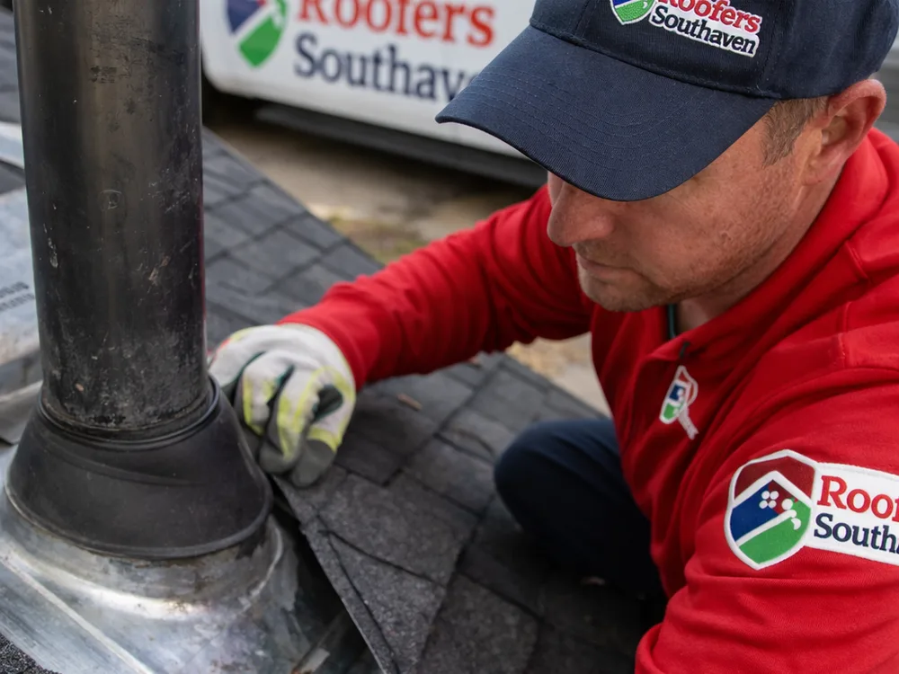 Roofer checking flashing and pipe boot condition near a vent stack on an asphalt shingle roof